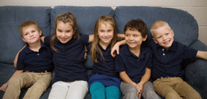 An image with 5 children sitting on a couch, with their arms around each other.