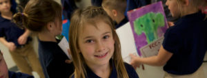 An image of a smiling girl, with other children carrying their artwork in the background.