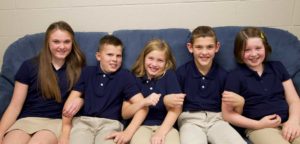 An image of 5 children sitting on a couch smiling, arms linked.
